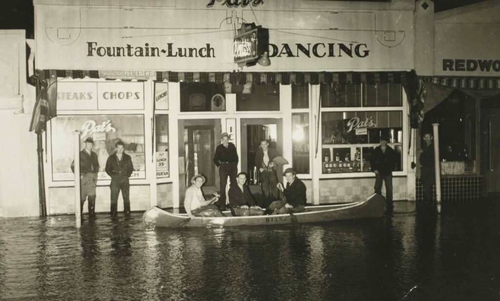 Flooded street in front of Pats Cafe, Guerneville, 1940. Three men paddling canoe and others standing at entrance. (Sonoma County Library)