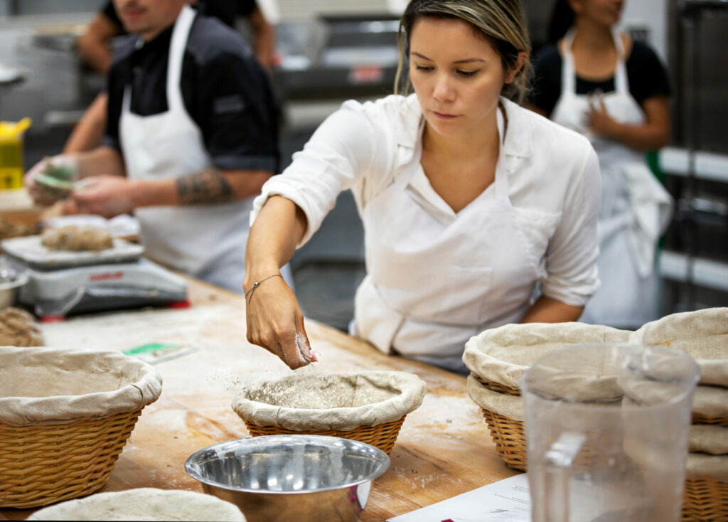 Instructor Pablo Puluke Giet teaching baking at the Artisan Baking Center Gabrielle Scrimshaw flouring her workspace before folding dough